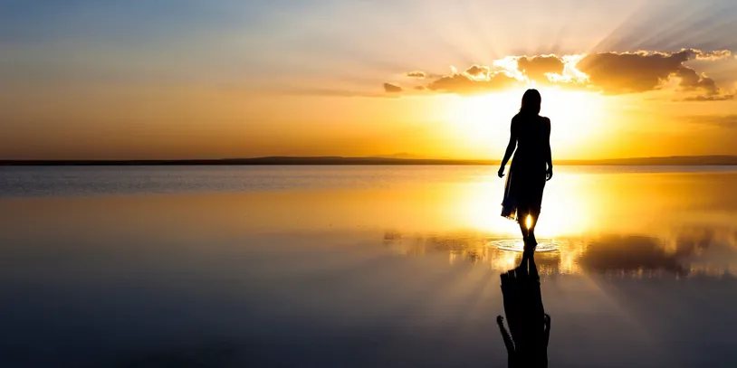 Silhouette of a person walking along the shoreline at sunrise with reflections on still water