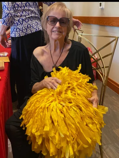 Portrait of Jeri smiling indoors while holding a bright yellow pom-pom